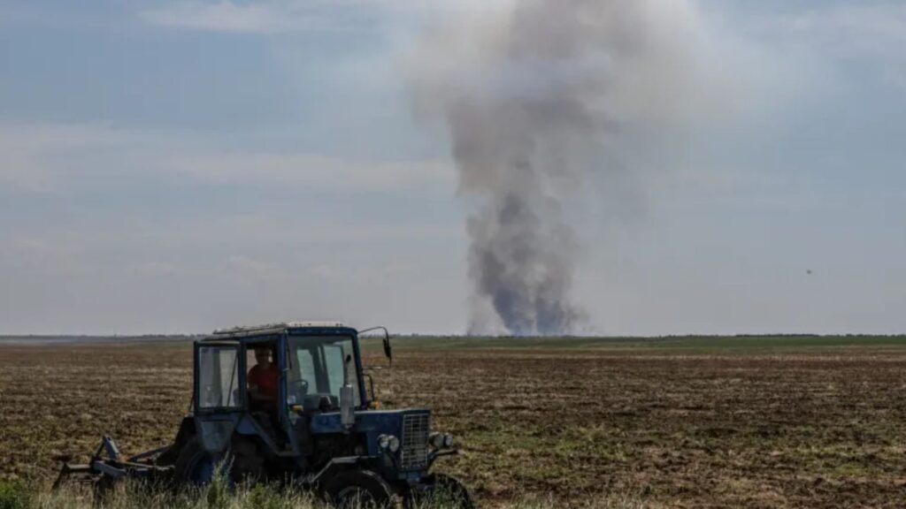 A farmer operates a tractor in the Kherson region as smoke rises in the distance after a military strike